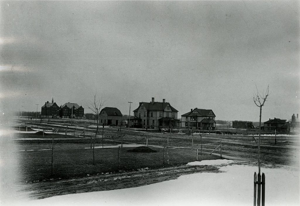 Macalester’s campus in the late 1800s. East and West Old Main are to the far right. There are houses in the foreground and a tree to the right.