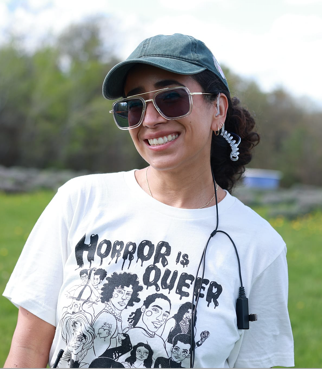 Photo of Emily Baeza smiling at the camera. She is in a green pasture, wearing a white shirt with horror characters which reads “Horror is Queer.”