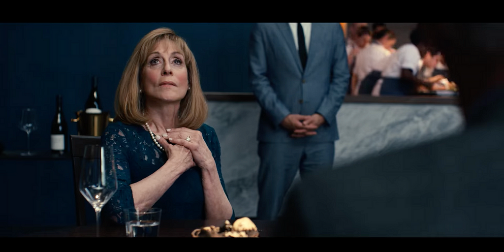 An image shows Judith Light as a character Anne Leiibrandt in a blue dress and short hair. She has a string of pearls around her neck and a wine glass in front of her on the table. She looks expectantly at someone off camera. She has a wedding ring on her left ring finger.