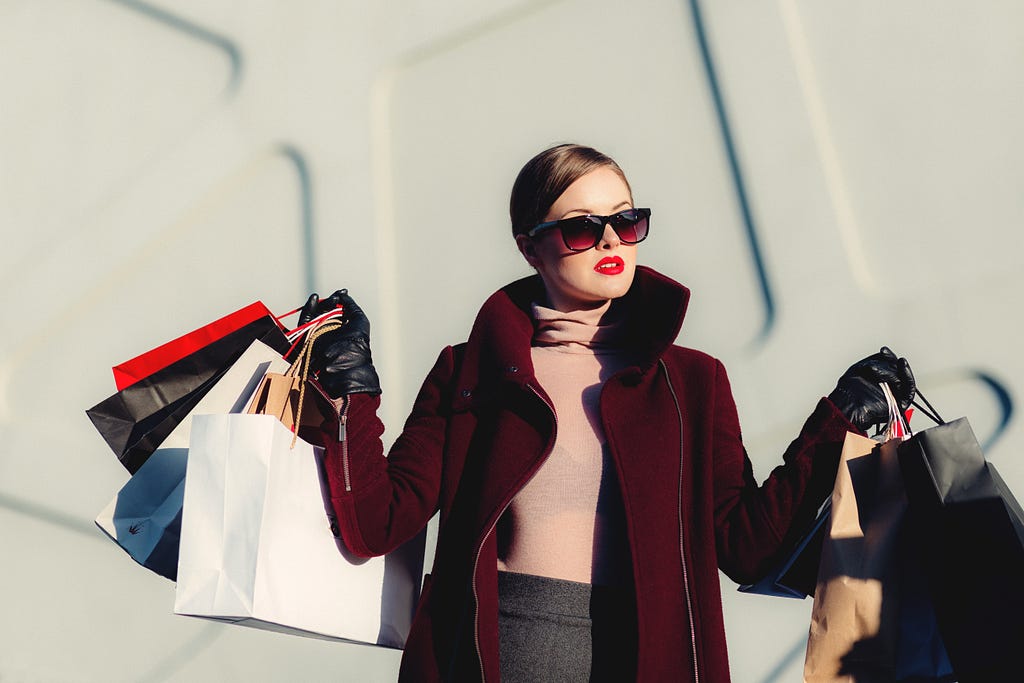 A white woman with blonde, tidy hair in a bun wears sunglasses and bright red lipstick. She hols dozens of shopping bags in both hands, her arms are outstretched to account for the weight of her items. The photo was taken during daylight.