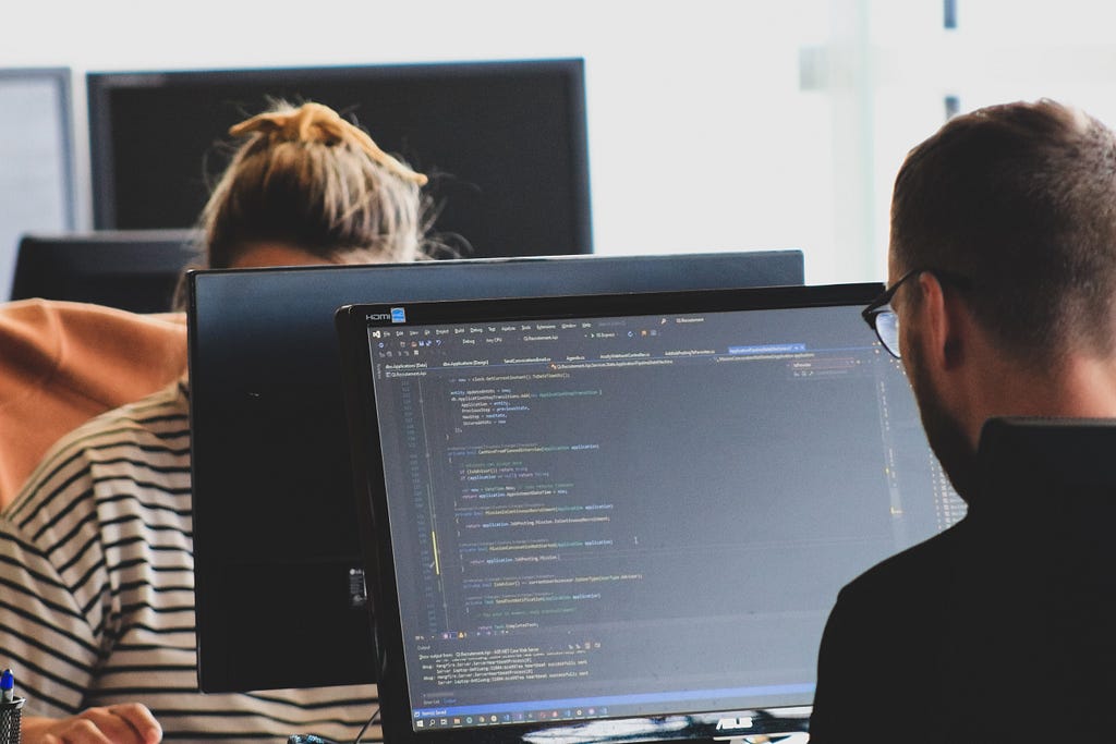 Two people looking at their own computer monitor, one with a text editor open for programming.