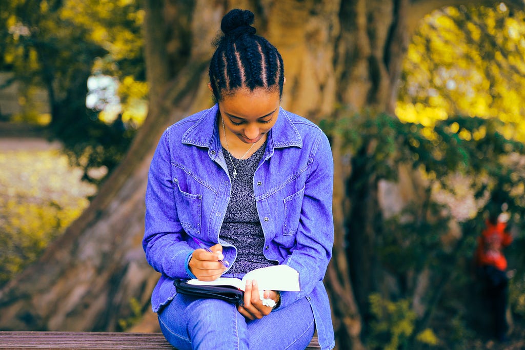 a young black girl sitting on a bench in a park with a book and pen in her hand. She appears to be quite interested in what she is reading