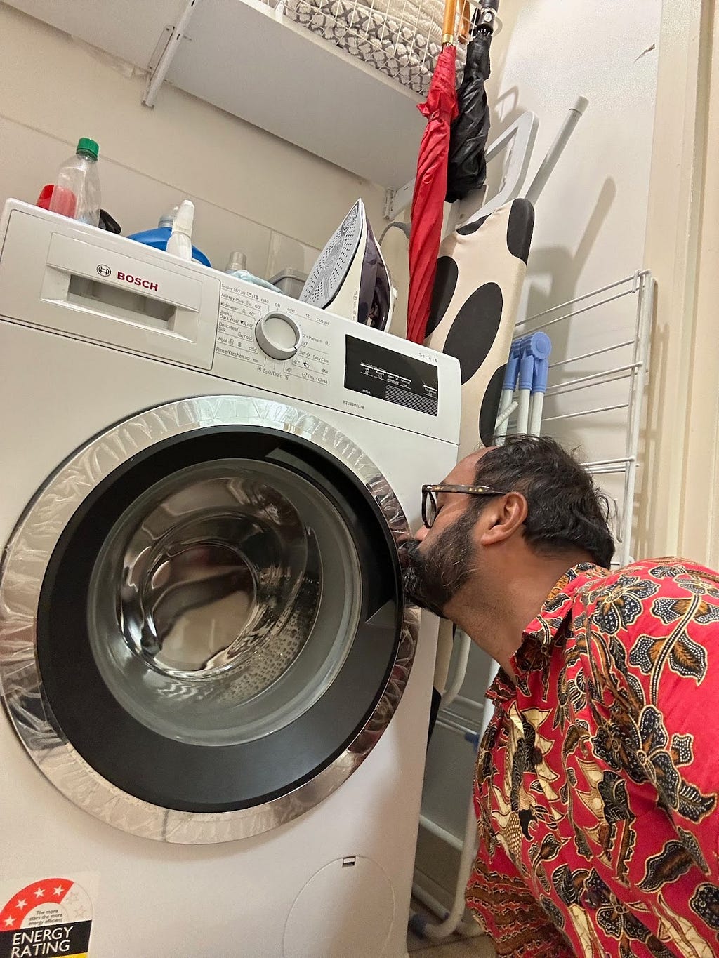 Brown man with glasses and red Batik patterned shirt kissing a washing machine.