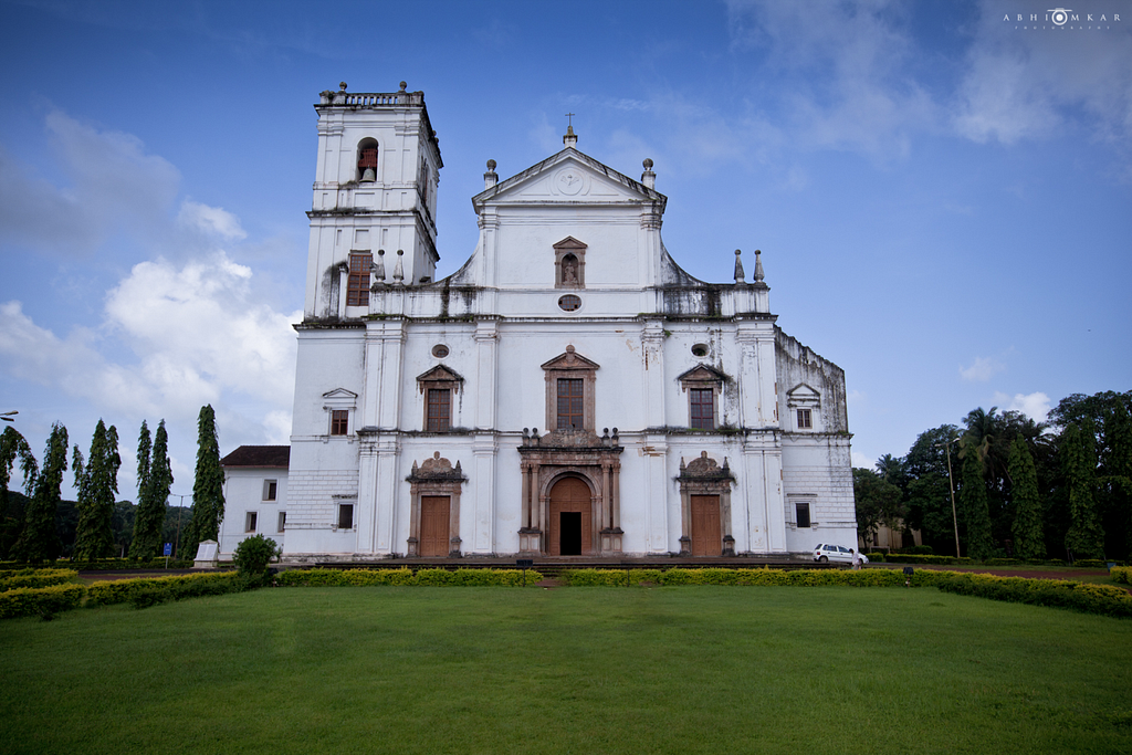 The Sé Catedral de Santa Catarina, a grand white colonial-era church in Goa, India. The façade features a mix of Baroque and Portuguese-Gothic architectural styles, with arched doorways, ornate details, and a bell tower. Lush greenery surrounds the historic structure.