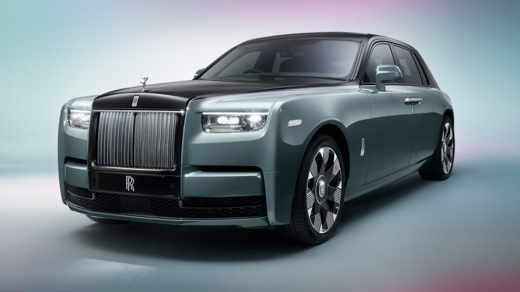 El nuevo Rolls-Royce Phantom está representado en una vista frontal de tres cuartos contra un fondo degradado de rosa a azul. El coche tiene una parrilla grande con lamas verticales y el adorno del capó Spirit of Ecstasy. La pintura es de un color verde azulado apagado, con líneas elegantes y ruedas negras pulidas con el emblema de Rolls-Royce en el centro.
