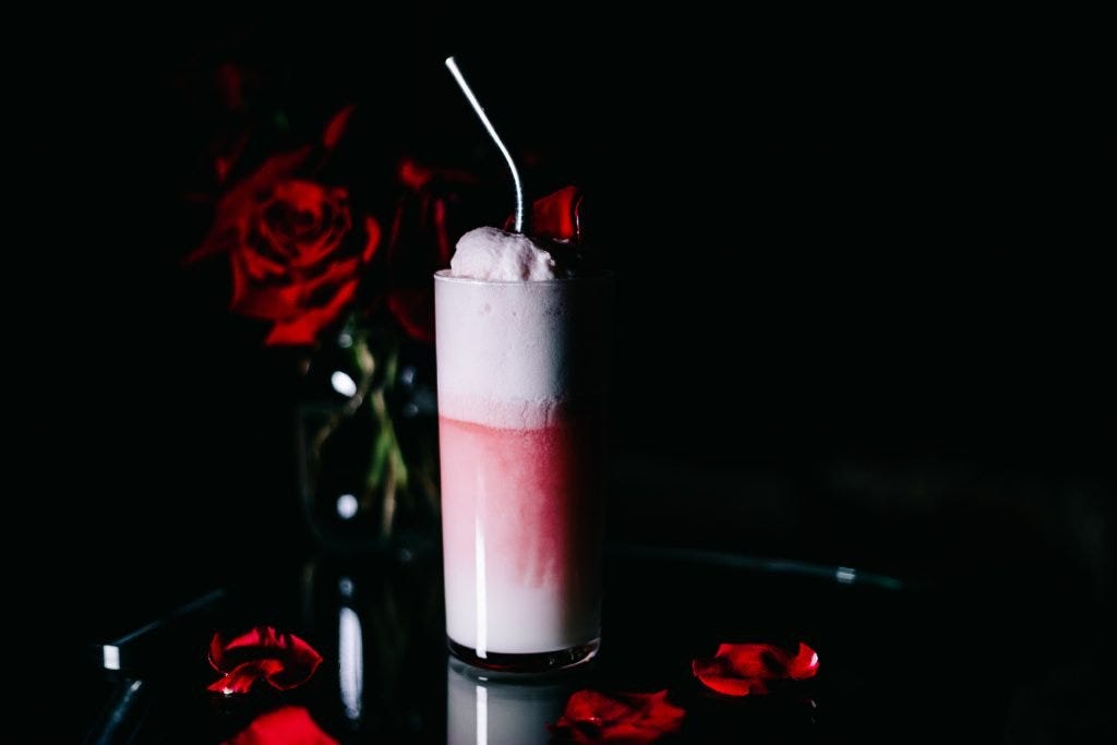 A pink cocktail with a silver straw sticking out the top. There are roses in the background and petals in the foreground