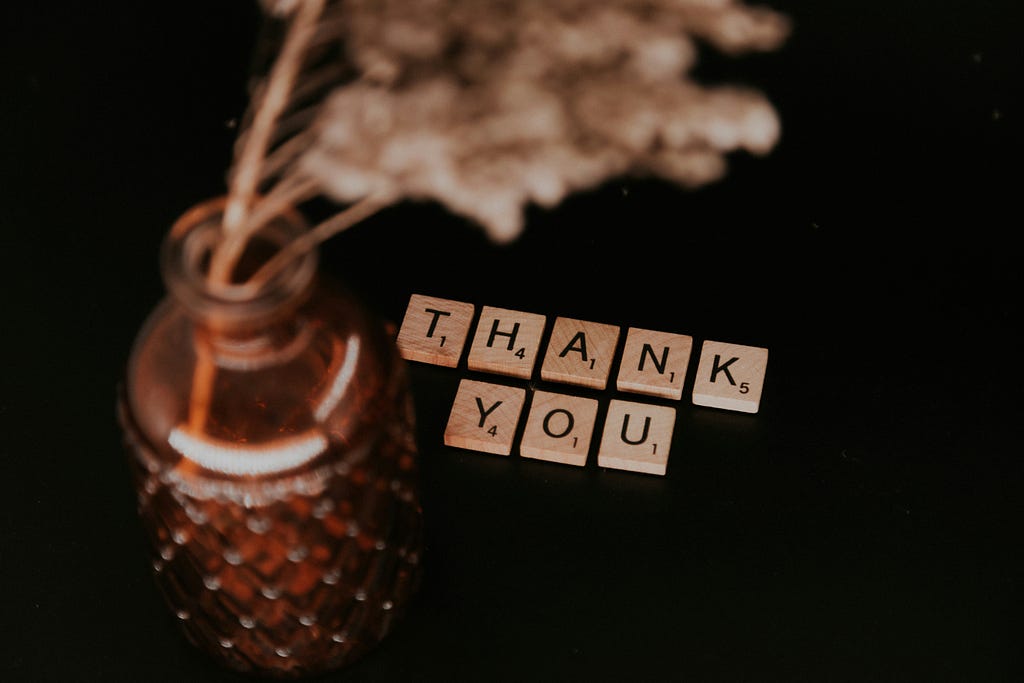 This image depicts a flower on a jar, with scrabble pieces on the side spelling out “thank” and “you.”