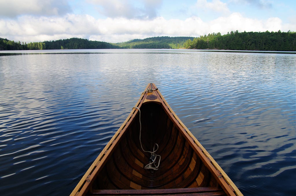 The front of a wooden canoe looking out over peaceful water and a green landscape in the distance