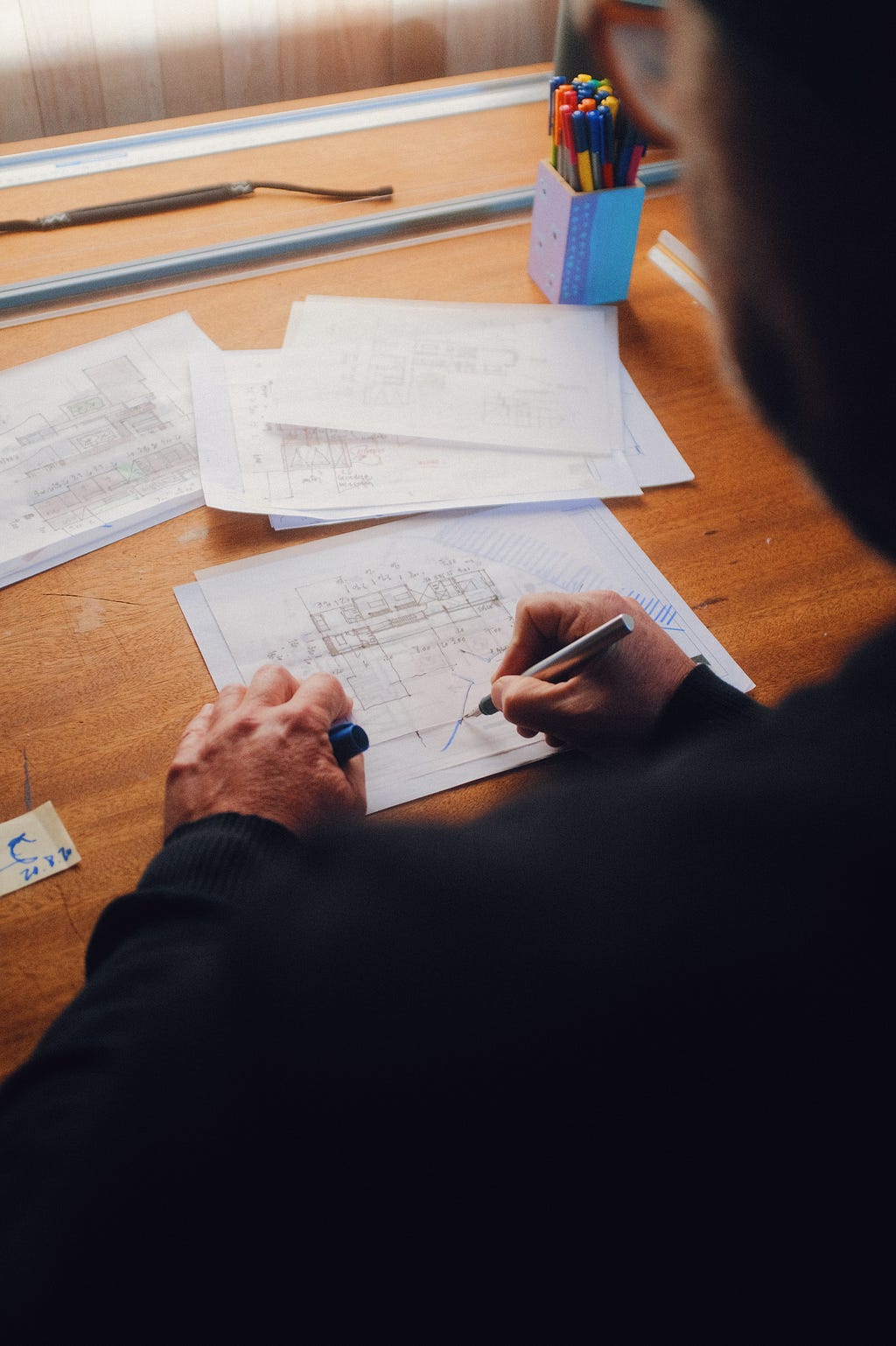 A photo of a desk with pieces of paper on it. One of the pieces is positioned in the center and is being worked on by a person wearing a black sweater and holding a pen in the right hand.
