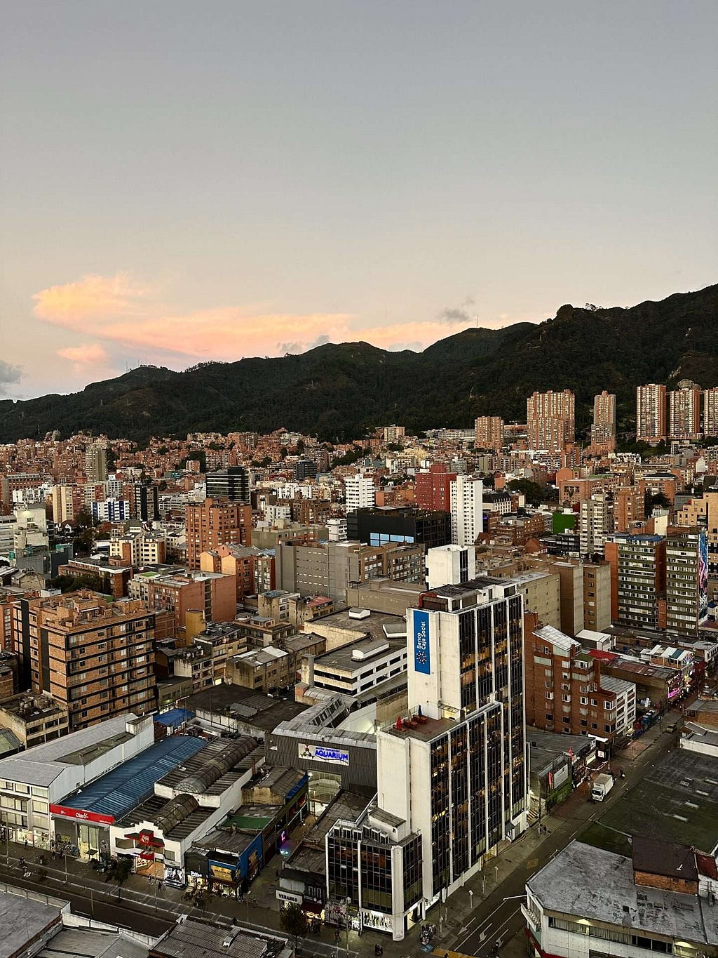 Aerial view of Bogota with numerous buildings, streets, and a backdrop of mountains during sunset.