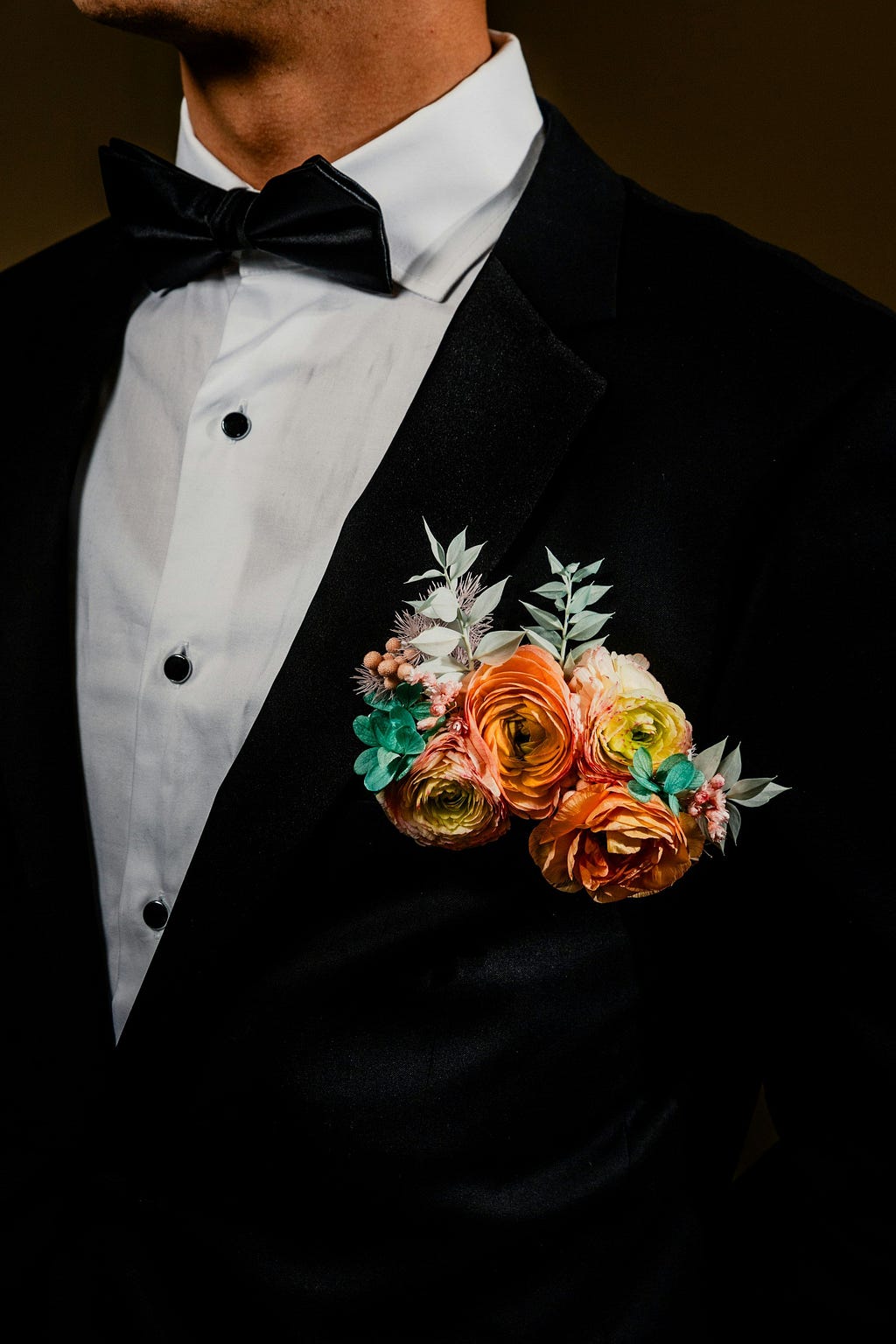 A formal black tuxedo with a white shirt and black bow tie, adorned with a colorful boutonniere featuring orange and peach flowers, against an indistinct background.