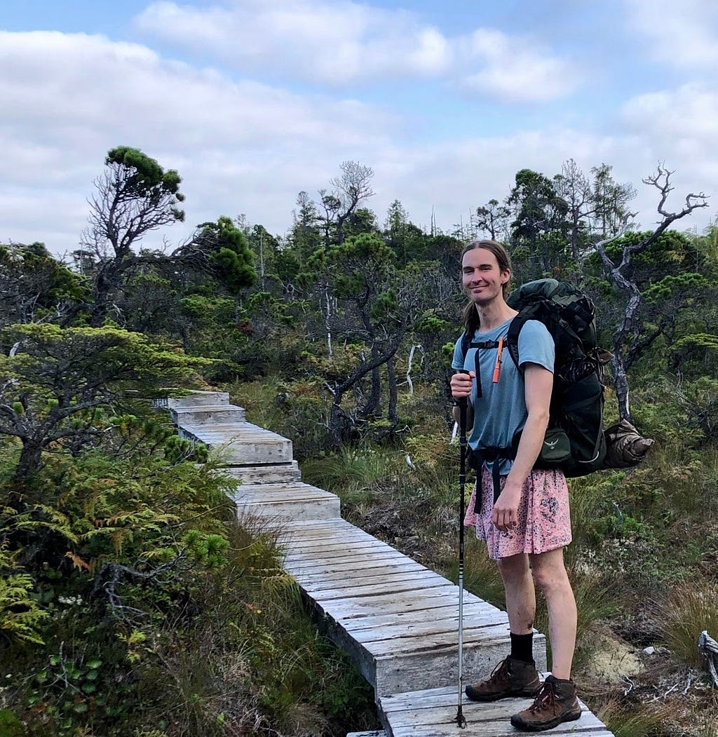 Myself: White 37 year old with pony tail, wearing pink skirt, blue t-shirt, backpack and hiking pole, on a wooden boardwalk in the trees on the top of small mountain. Blue sky and clouds.