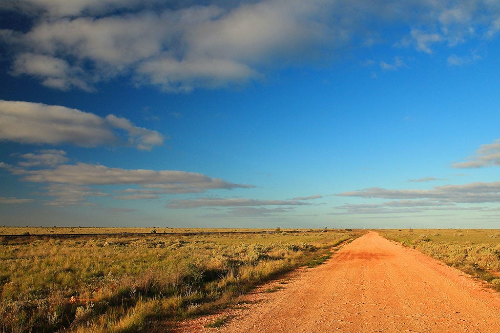 A red dirt road stretches into the horizon of the Nullarbor Plain under a vast sky with scattered clouds.