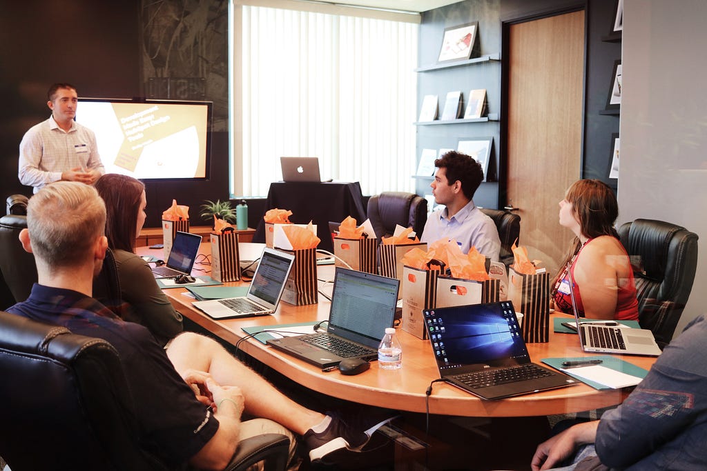 Team of 5 people sitting in an office board room with laptops, doing training with a presenter.