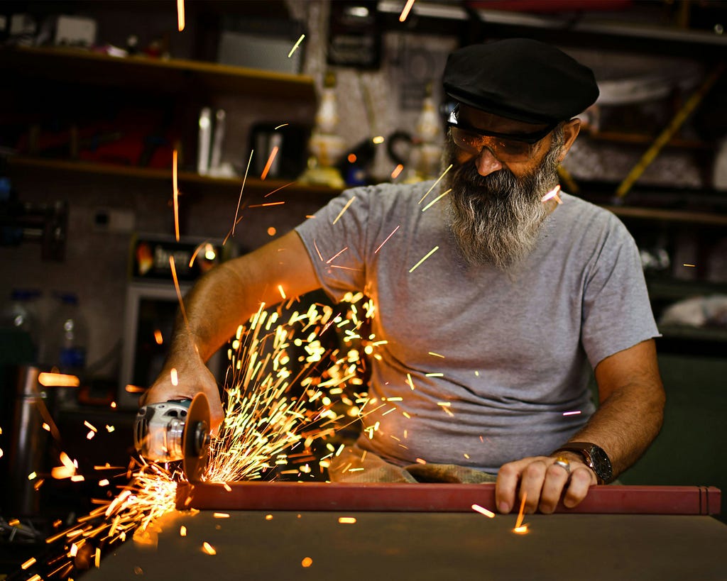 A man with gray and black beard wearing a short-sleeved cotton gray shirt, hat and protective goggles uses an electric circular blade to cut through a section of 2-inch steel beam as sparks fly.