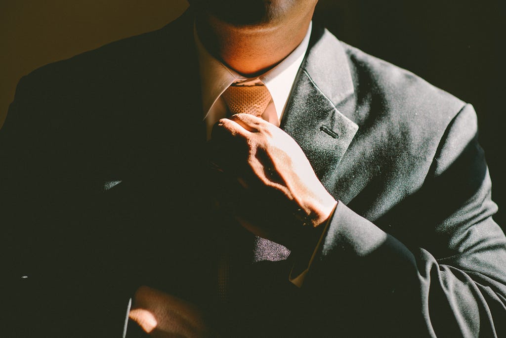 Photo of a man wearing a grey suit and adjusting his tie. The photo has been cropped to show just his chest and neck.