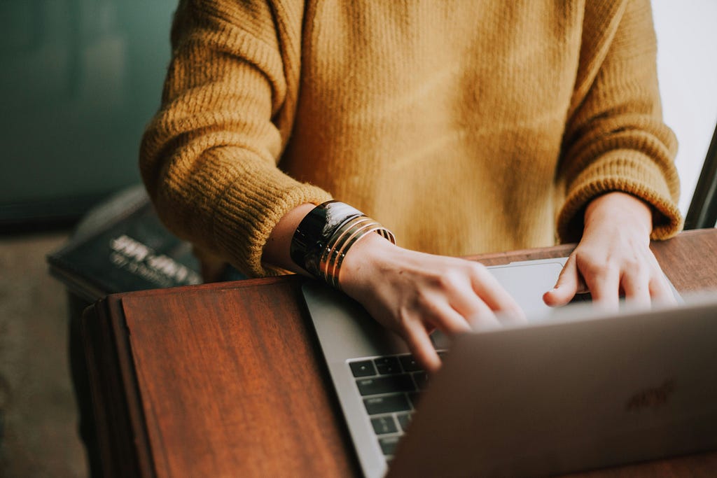 person wearing an orange sweater sitting at a wooden table typing on a laptop