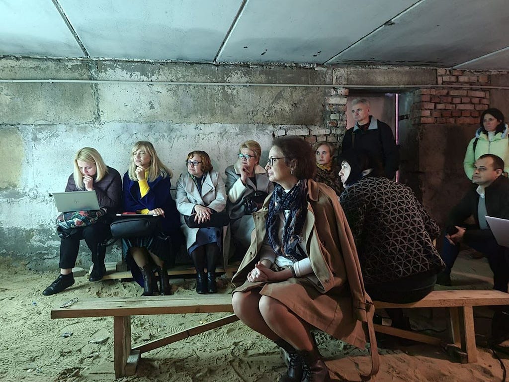 A group of people wearing coats and scarves and sitting on benches while holding a meeting in an underground bunker in Ukraine.