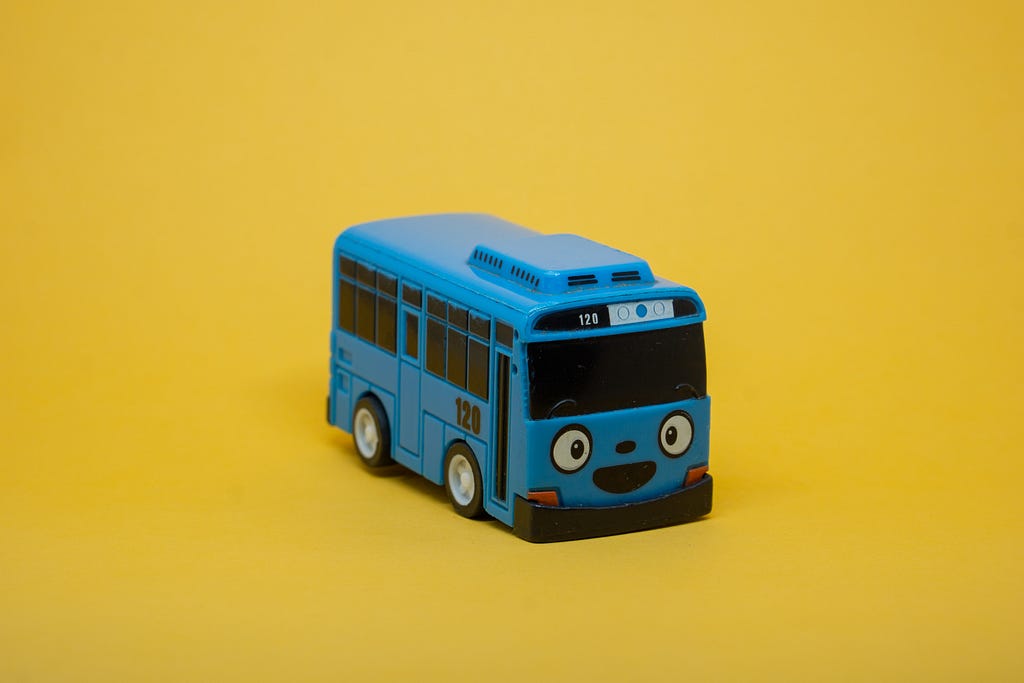 Small bus toy on a bright yellow background. The bus is blue, with wide eyes and a big, black smile.