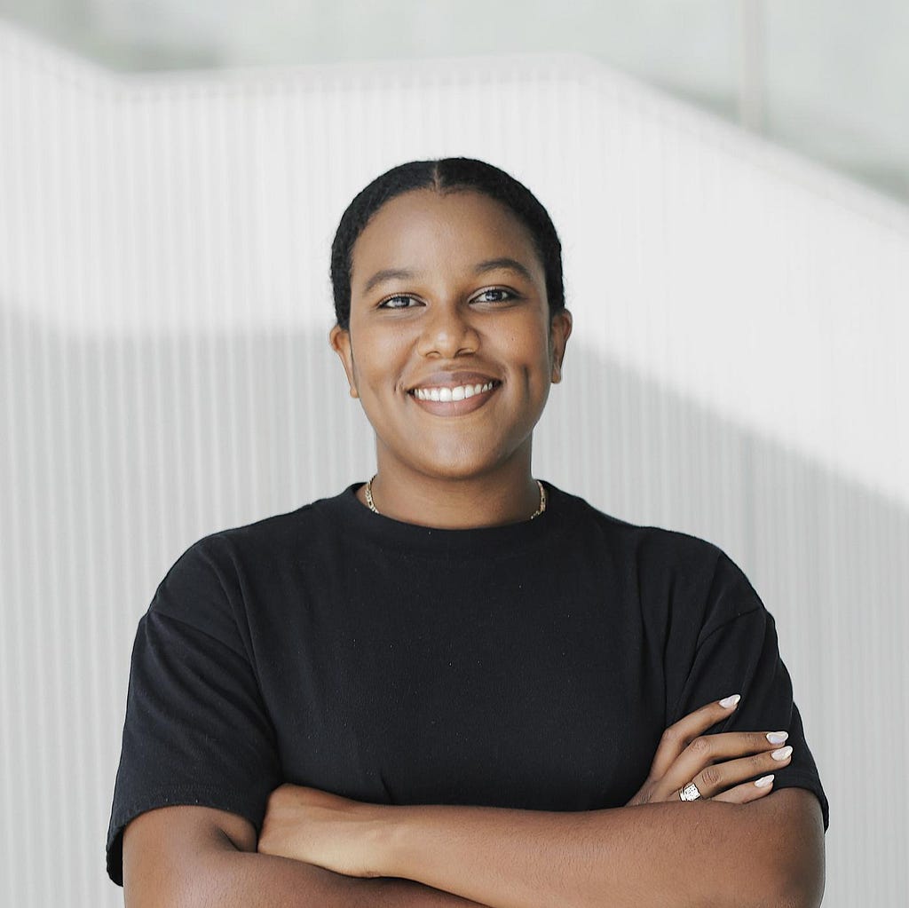 A woman in a black shirt stands in front of a white background and smiles directly into the camera.