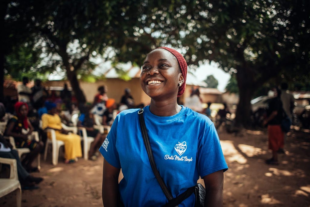 Photo of Ruth Alanana, a 25-year old “Data Advocate” for the Data Made Simple project. Ruth is standing outside wearing a blue t-shirt with text “Data Made Simple.” Ruth is standing outside in a rural setting of Nigeria, smiling, and looking off camera. In the background are the backs of people seated in rows.