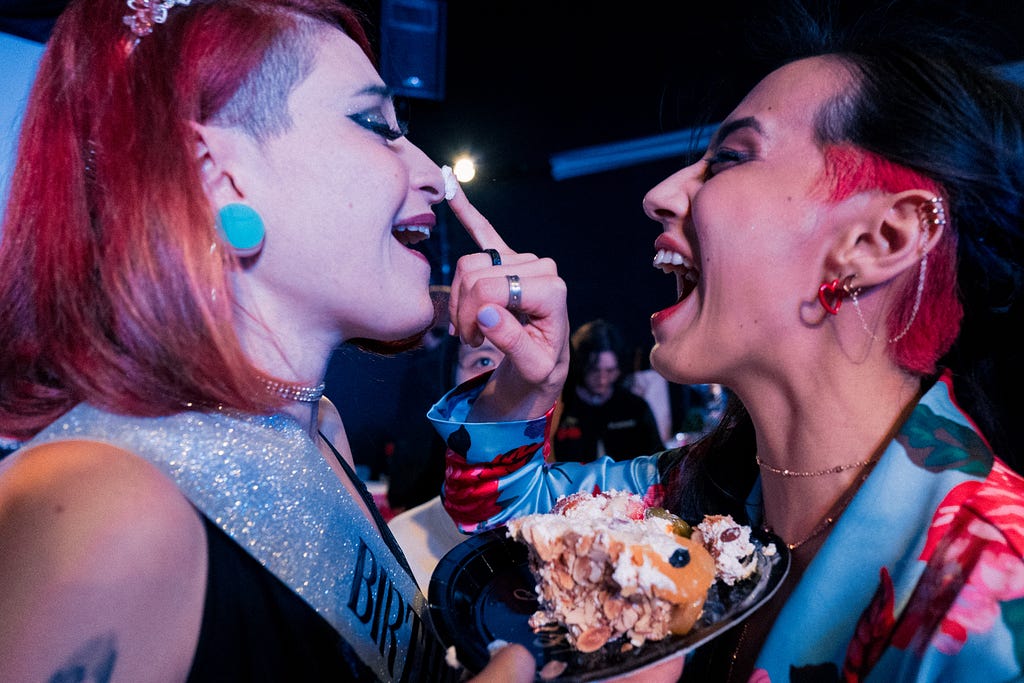 Two makeup laden lesbians putting cake frosting on each others noses and laughing