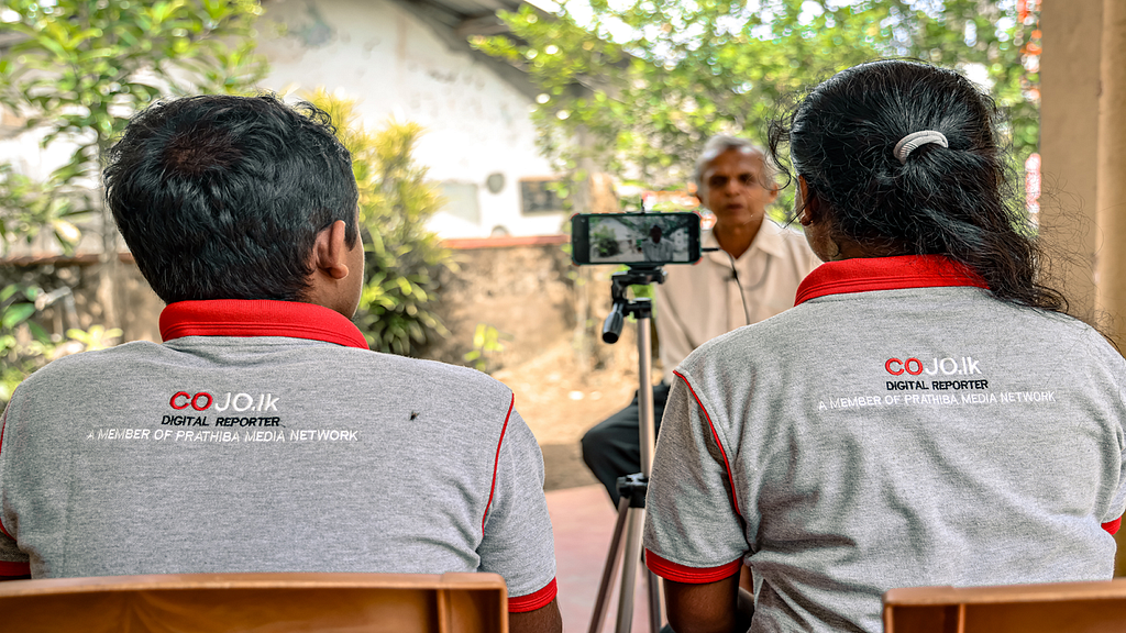 Two people wearing matching branded shirts that identify them as digital reporters sit side-by-side with their backs to the camera while interviewing a gray-haired man with a mobile phone perched on a tripod.