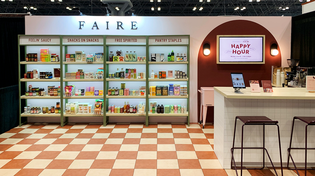 Photograph of Faire’s Summer Fancy Food booth with four shelving units of products, a checkerboard print floor, and a small cafe-style seating area.