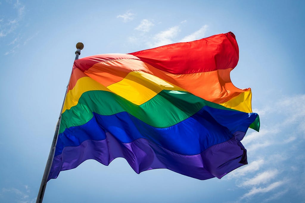 A flag with red, orange, yellow, green, blue and purple colours against a blue sky. A rainbow flag usually carried by members of the LGBTQIA+ community.