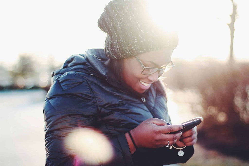 Dark skinned woman walking in cold weather checking her smartphone, smiling and scrolling