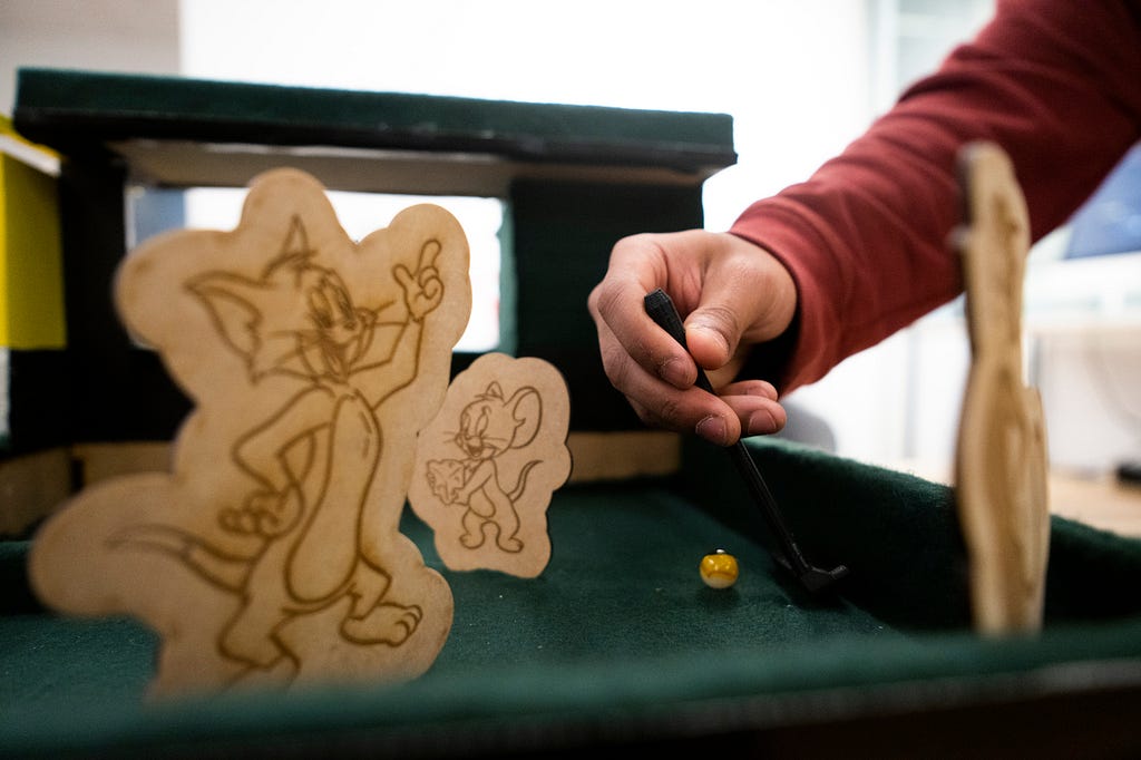 close-up of person’s hand using hand-held putter to tap a marble around laser-cut certical obstacles of Tom and Jerry