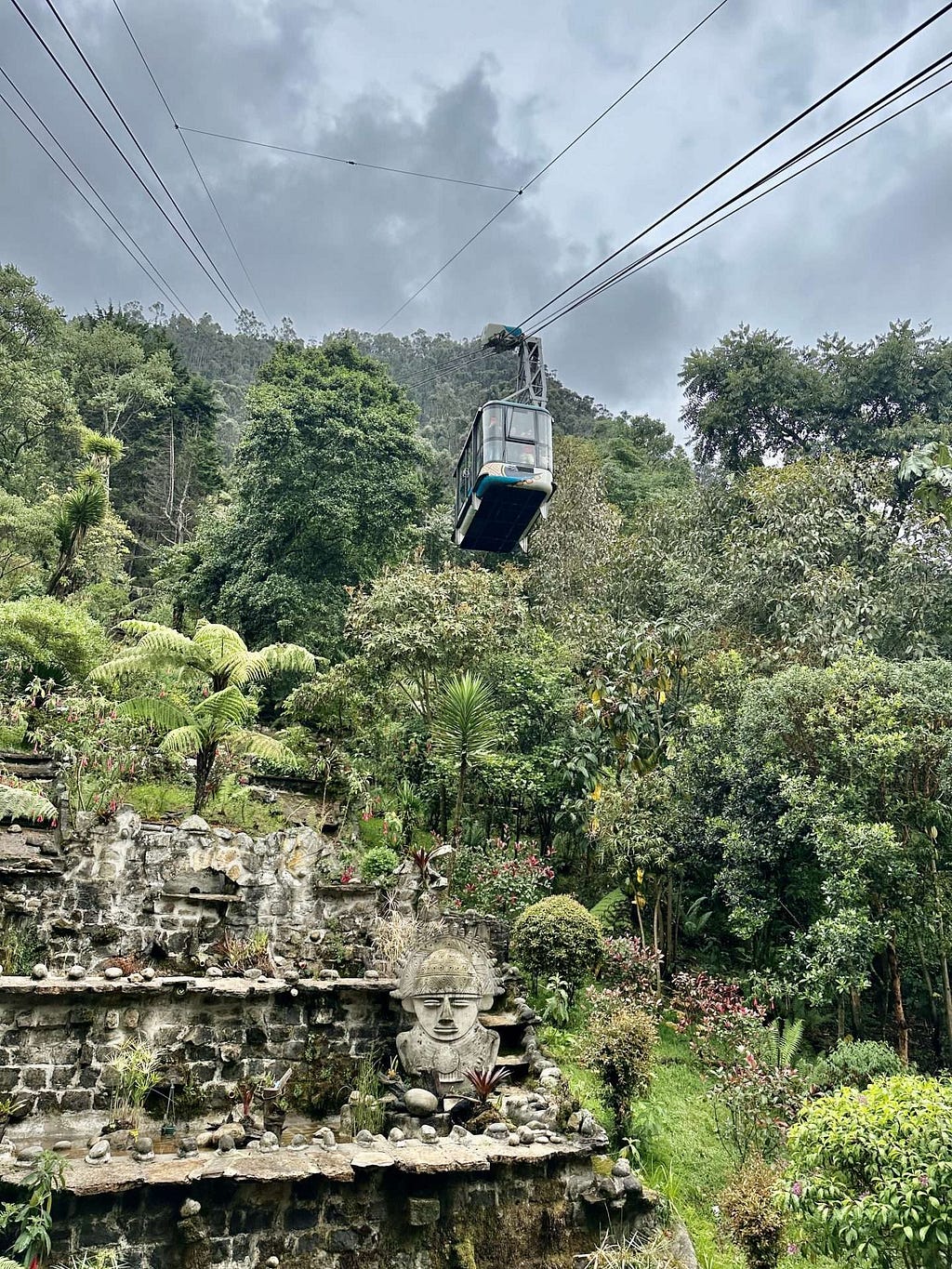 A cable car travels over a lush, green hillside with a stone terrace below featuring a carved face statue. Dark clouds loom overhead.