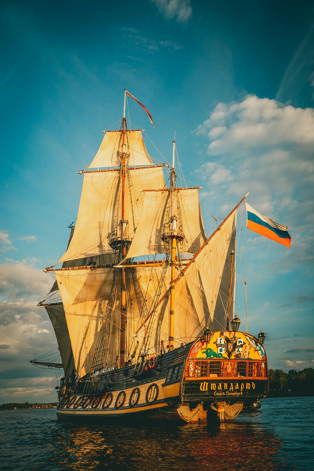 A classic tall ship or galleon with multiple sails, sailing at sunset with a flag featuring horizontal stripes