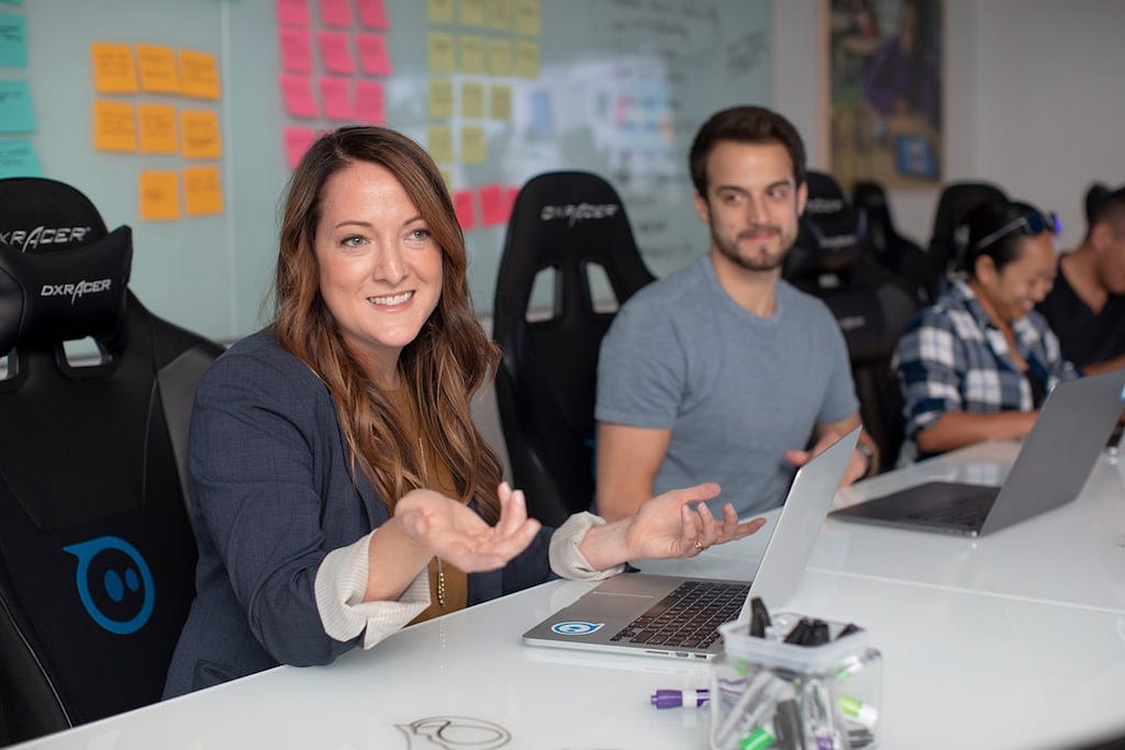 A team of professionals are having a meeting, laptops are on the table and a young woman is talking while her colleague looks on.