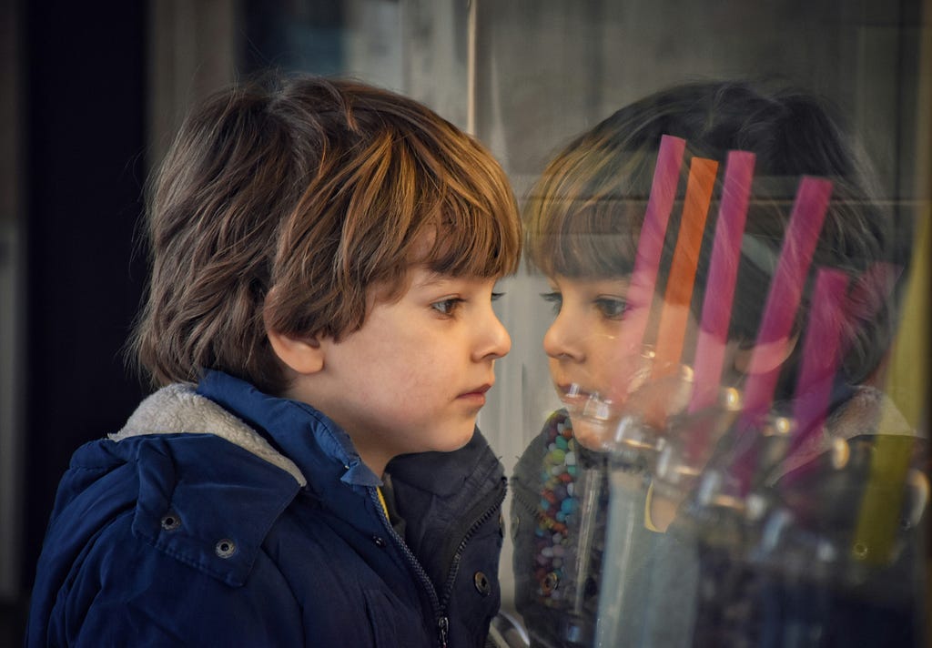 A child staring pensively at his own reflection in glass
