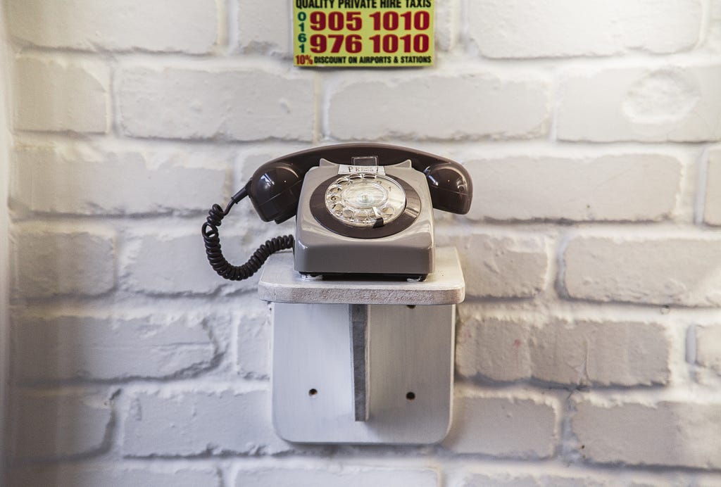 A grey telephone sits on a shelf in front of a brick wall. A card advertising a taxi firm number is above it