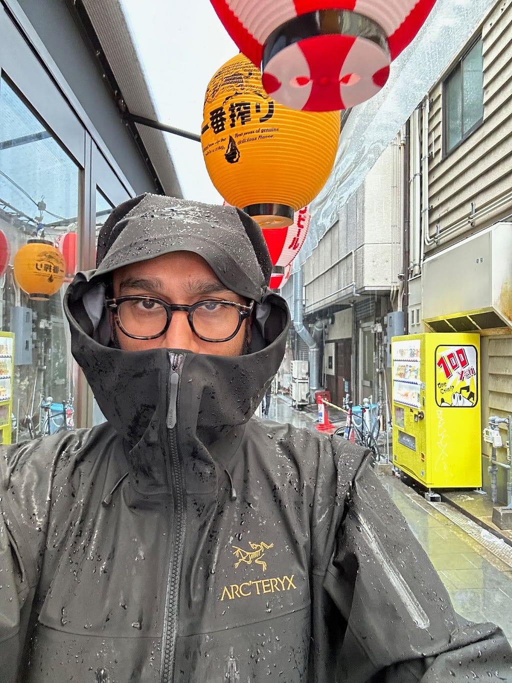 Selfie of brown man with glasses standing in rain in an Arc’teryx outer shell with hood drawn up. Japanese signs, lanterns and vending machine in the background. Wet street.
