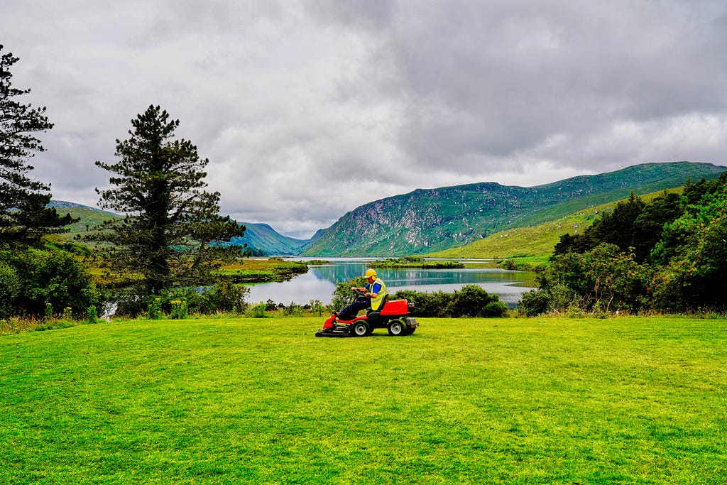 A man on a riding lawnmower over lush grass with a lake and mountain in the background