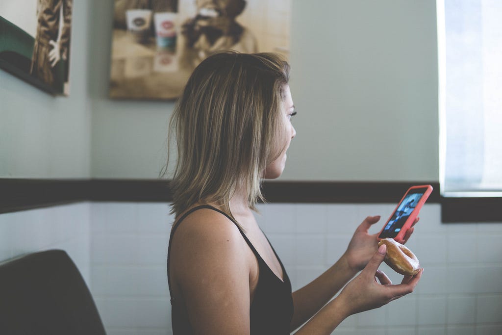 A woman glances at her smartphone while holding a donut.