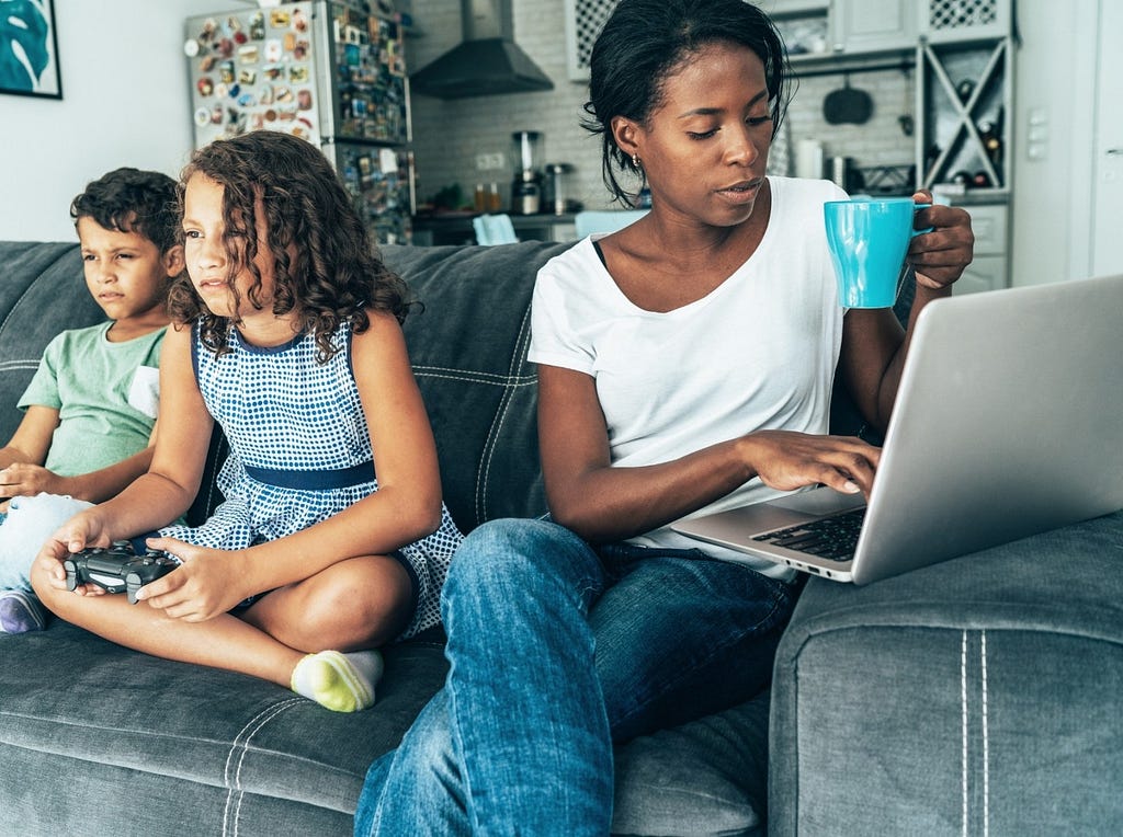 Woman works on a laptop while her children play video games. Photo by filadendron/Getty Images