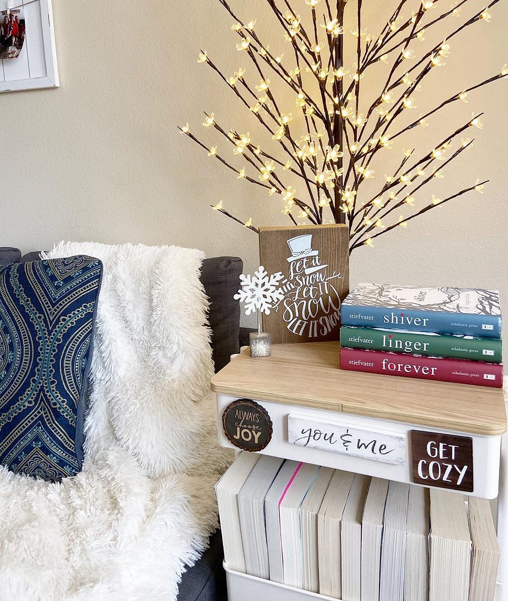 An accent chair with a white throw blanket and decorative pillow next to a cart with home decor accessories and books.