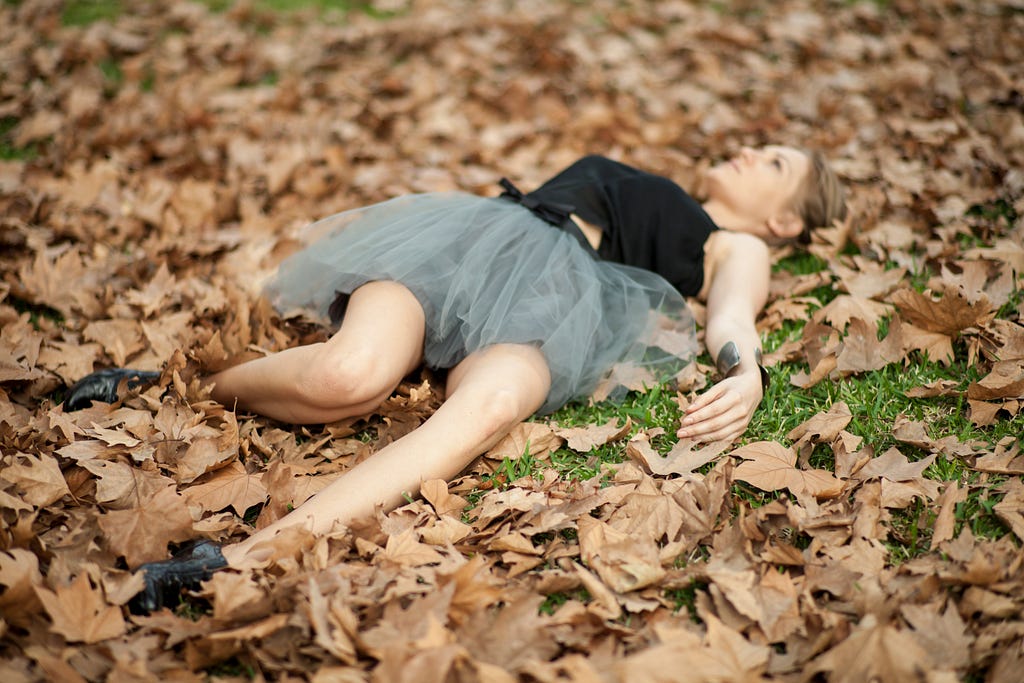 woman in a black shirt and grey tulle skirt layong on the a bed of orange autumn leaves with knees and limbs splayed around like she fell.