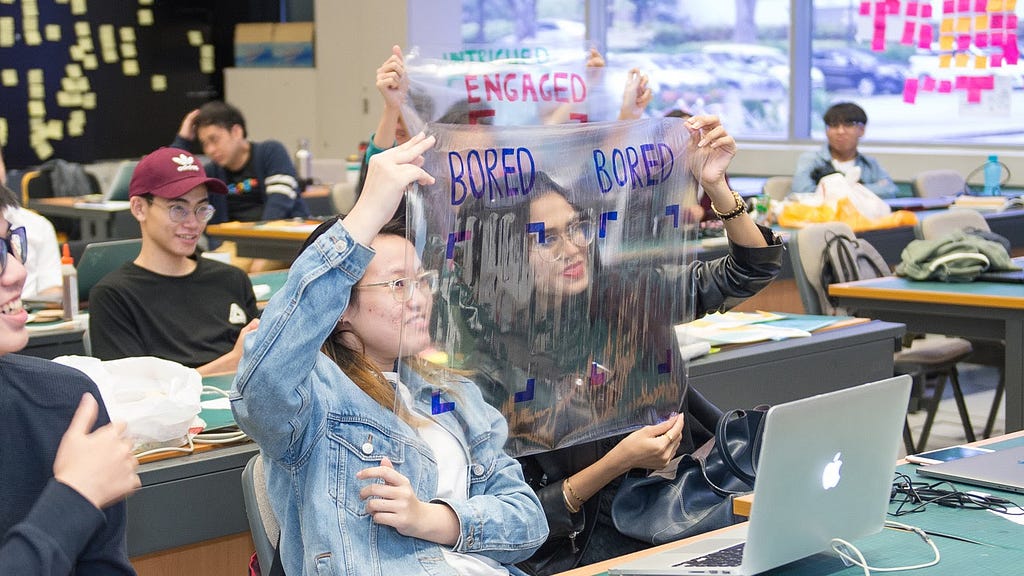 A classroom of students demonstrating an AI prototype. They are holding clear acetate sheets in front of their faces. Each sheet has a drawing of a frame with words above it — bored, engaged, intrigued.