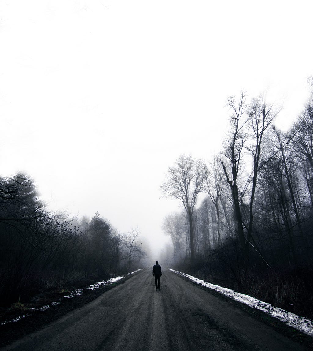 A man walking alone on a road in between two sides of dark forest on a cold winters day. Photo is in black and white.