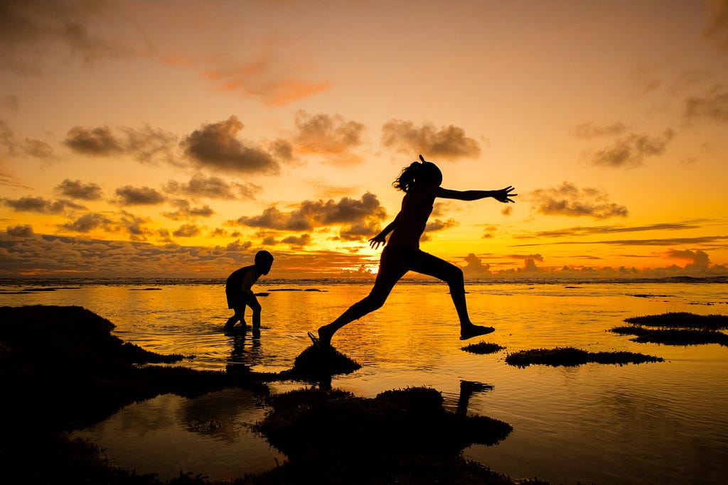 photo of two children playing on the shore, during sunset. The silhouettes of the children show one jumping to the water, and the other standing in the shallow water, with small mounds peaking from the water’s surface, maybe grass, rock, or sand. The sunset behind them is shades of golden yellow, orange and pink, dotted with fluffy clouds.