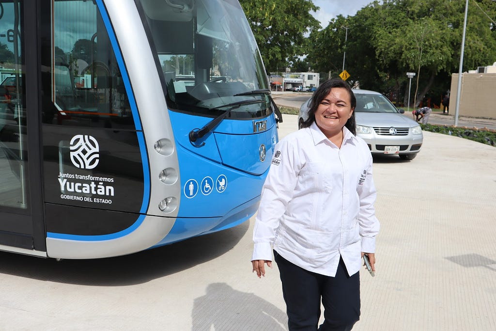 A smiling woman stands in front of a blue and silver bus with a silver vehicle coming into view over her left shoulder.