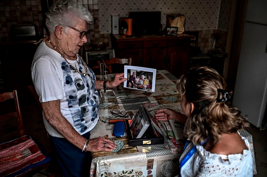 This is a photo taken at someone’s home. A young person is sat, back to the camera, looking at the photos an older, standing camera facing person is showing them. They appear to be inside the older person’s dinning room. The scene is set around a table with a waxed cloth and trinkets on the furtniture in the background. The light is dimmed, as they might have closed the curtains during the day to prevent sunlight from coming in.