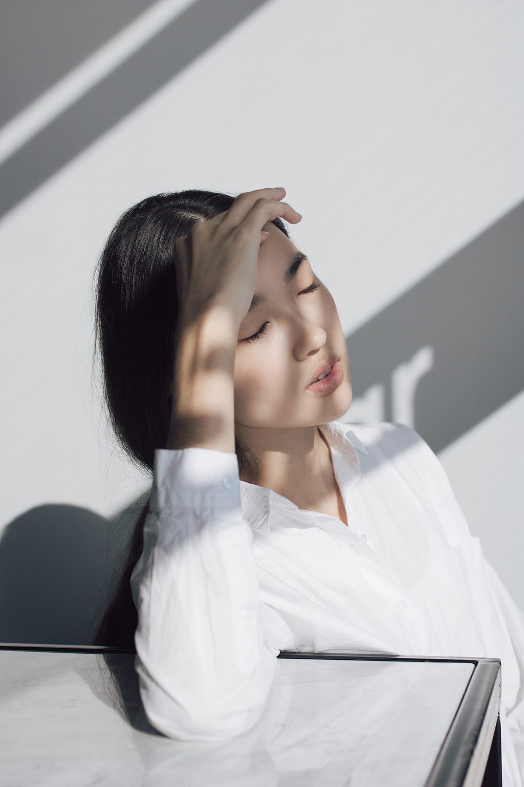 Fair-skinned young woman with neck-length straight, black hair and wearing long-sleeved white blouse. Her partially shadowed face, with eyes closed and lips partly opened, is upturned slightly sideways to the right, her right arm resting on a counter top and right hand on her forehead, as if massaging it.