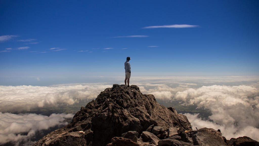 Um homem de boné está estático sobre um pico de uma montanha, observando o horizonte com nuvens.