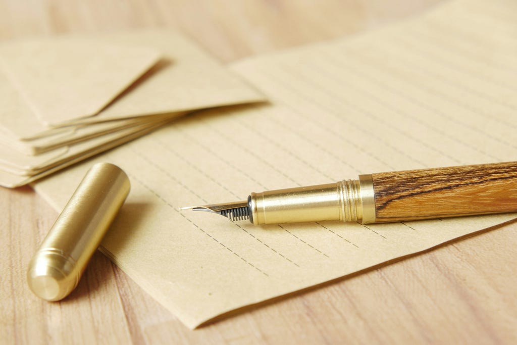 Paper underneath a wooden fountain pen with a gold top, envelopes in the upper left.